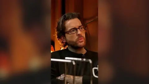 Pete wicks sitting down with his arms crossed 