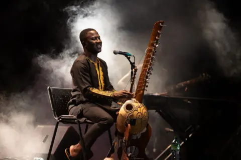 JEROME FAVRE/EPA Smoke is in the background as Lamine Cissoko smiles whilst he plays the kora.
