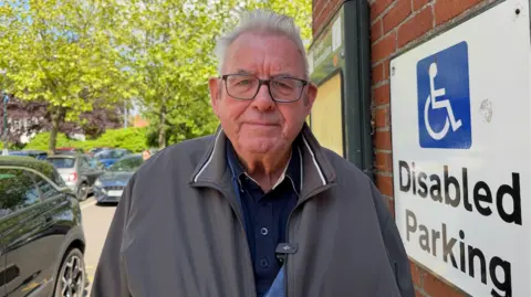 Alan Ward is in a car park and is wearing a grey jacket with a navy top underneath. He has glasses and white hair. He is standing next to a disabled parking sign.