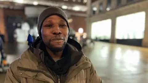 Josh looks into the camera. He is wearing a black hat and a grey-green coat over the top of a black hoody. Behind him, out-of-focus, are shops in the centre of Leeds