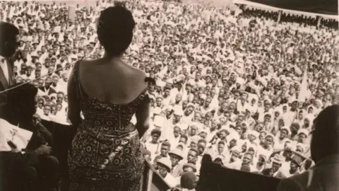 Herbert Weiss Wearing a plaid dress, Andre addresses a large crowd of men and women at a rally in the Democratic Republic of the Congo