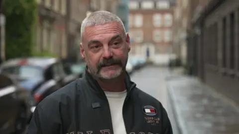 Chris Lloyd mid-interview. He has short white and grey hair and a short beard. Elder Street is in the blurred background, with brick buildings and some parked cars along the right-hand side.
