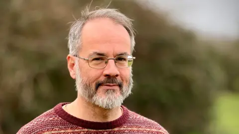 A man stands outside with a large hedge blurred in the background. He is wearing wire rimmed glasses, has short grey hair and a beard. He is wearing a patterned red jumper.