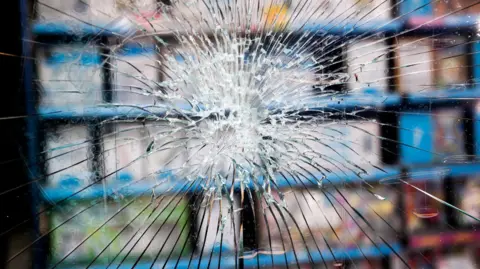 A smashed window of a shop, with the goods seen out of focus behind it. The glass has been smashed into shards but has not fallen out of the window.