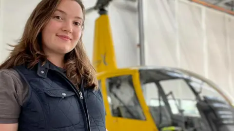 OLIVIA WATSON Olivia, who had long brown hair and is wearing a blue sleeveless zip-up top and grey T-shirt, standing next to a yellow helicopter in a hangar.