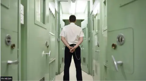 A prison officer, wearing a white shirt and black trousers, stands with his back to the camera. There are pale green prison doors to the left and right of him