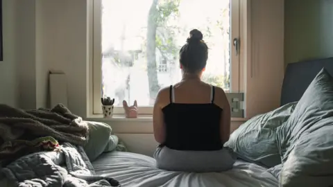 Getty Images young woman sitting in bedroom facing a window with her back to the camera