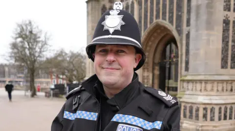 Shaun Whitmore/BBC Sgt Chris Clay. A man who is looking at the camera and smiling. He is a police officer who is dressed in a black police uniform with a coat. He is also wearing a police hat and is standing outside a church. 