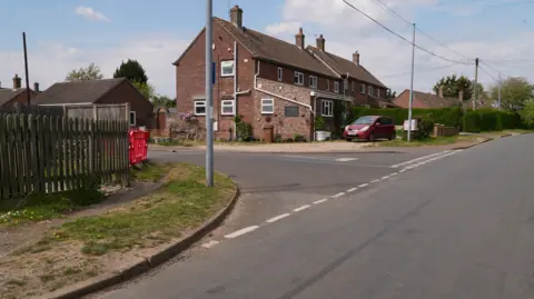 Martin Giles/BBC A street view shows a junction at a road. There are redbrick houses and cars parked on driveways, with a long skid mark visible on the road surface.