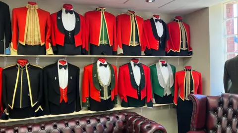 The tops halves of dress uniforms are seen on two shelves on a wall. The jackets are mostly red, some are black, and they have varying gold trims and some with black or green cloth underneath, some with high collars and others with bow ties at the collar.