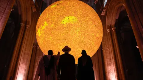 A big sphere illuminated from within in yellow, suspended inside a cathedral with three people standing in front of it in silhouette.