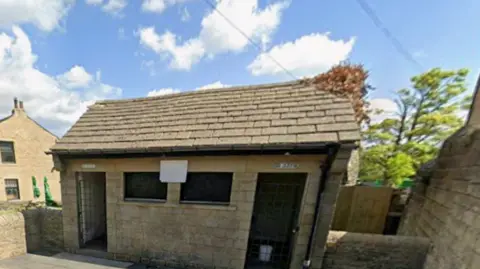 The image shows a small, single‑storey stone building with a pitched roof of grey stone slates and a dark gutter running along the front edge. There are two dark, rectangular window openings. At either end are doorways. Above each doorway are small signs, 'Ladies' and 'Gents'.
