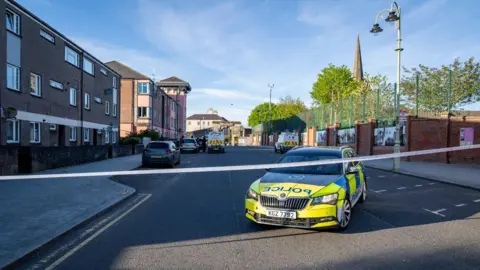 Aodhán Roberts A police car on a street behind tape which has the area cordoned off. Houses are in the background at the left and a spire, trees and fence are on the right.