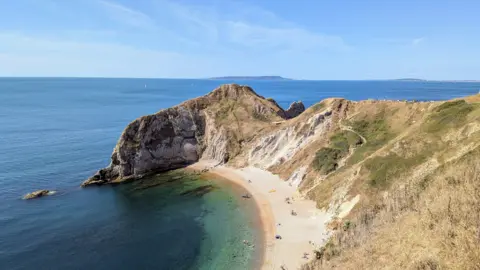 Paul Horsley Mountainous cliffs tower over a small isolated beach. Behind the cliffs we can see the line of the horizon.