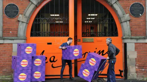 Visit Worcestershire Purple boxes of Worcester sauce Walkers crisps being taken into the large orange doors of the Lea & Perrins site. One man is carrying a box and another has two boxes on a trolley. Black plaques with gold lettering flank each side of the door.