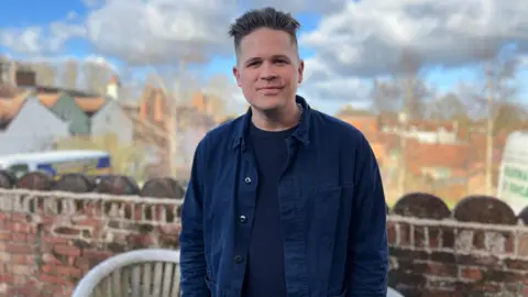 Jon Wright/BBC Luke Wright stands in a garden with houses and trees in the background