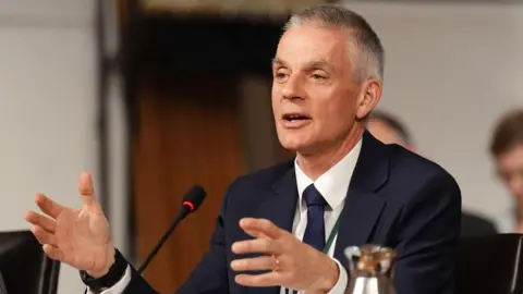 Picture of Tim Davie from March 2026 in a blue suit with a white shirt and a matching tie