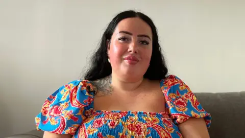 Suzie is seated on a sofa wearing a bright blue dress with a red and yellow floral pattern. A small tattoo is visible near her shoulder, with cushions and a plain wall in the background.