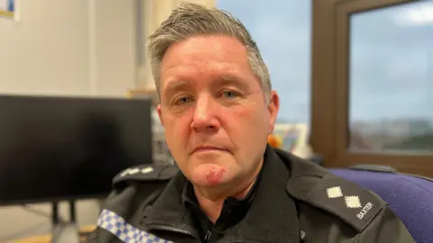 A police officer wearing his black uniform is sitting in an office with a black computer screen and windows behind him. He has two stars on his shoulder and the word "Baxter". He has blue hair and grey eyes.