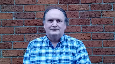 a man with combed-back dark hair, in a checked shirt, standing with a red brick wall behind him