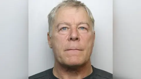 Headshot of a man with grey hair and blue eyes. He is wearing a black shirt. 