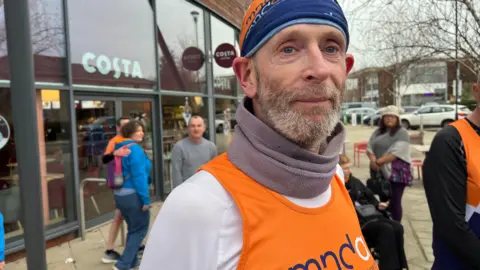 Anthony Cox wearing an orange high vis MND vest and a white top underneath it. He also has an orange MND head band on and has a light facial hair. There are people milling in the background, in front of a Costa Coffee shop.
