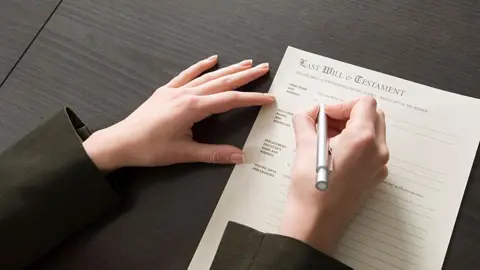 A person is filling out a 'Last Will & Testament' form on a dark wooden table. The person's left hand is holding the paper steady while the right hand writes with a silver pen. The form includes sections for 'Name,' 'Address,' 'Beneficiaries,' and other relevant details. The person is wearing a dark green jacket.