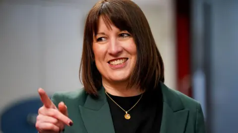 Reuters Rachel Reeves, with brown hair cut in a bob, points and smiles. She is photographed from the chest up and wearing a black top and green jacket. She is also wearing a gold pendant.