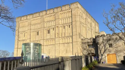 Norwich Castle keep, a square building with battlements and flagpole, built from light-coloured stone, set against a bright blue sky. A cylindrical lift can be seen in front of the castle, along with a ramp lined by railings. A tree with no leaves is on the right of the picture.