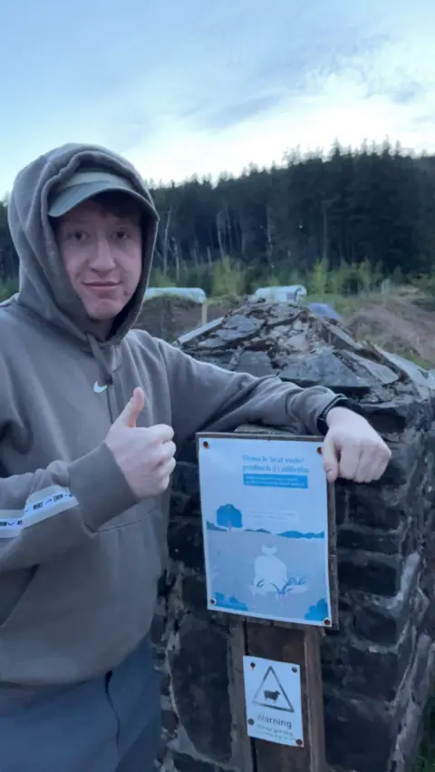 Family handout Charlie Owen is leaning on an information board relating to the woodland area he is in, posing with his thumbs up and looking at the camera in a hoody. Behind him are very tall trees.