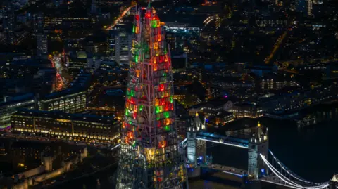 Shot from a helicopter at night, green, red and and orange lights shine in the top of the Shard with Tower Bridge in the background