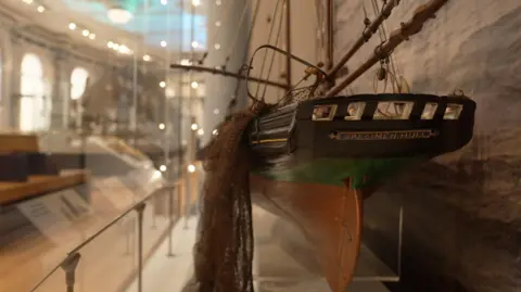 A small model of a boat is situated inside a glass case, and has "Specimen Hull" on the back.