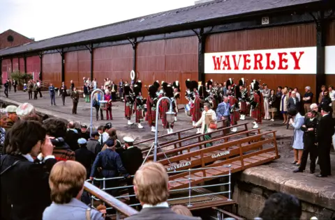PSPS A crown gathers on the Waverley as we look across to the quayside where a pipe band is playing to see it off. A well-dressed woman is walking across the passerelle and onto the boat.