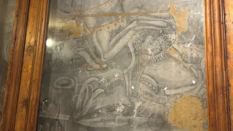 A faded mural on a wall, with wooden frames around it, showing a man on the ground and a horse. The image is hard to make out but is mostly grey and brown, with bits missing.