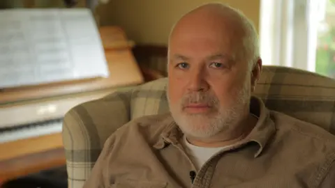 A man with shaved head and short beard loosk straight at the camer. He is wearing a brown shirt and is in an armchair. A piano can be seen in the background.
