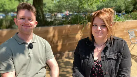 Cameron in a light green t shirt to the left, and Tash with a dark denim jacket, smile while sitting under the shade of a tree on a sunny terrace