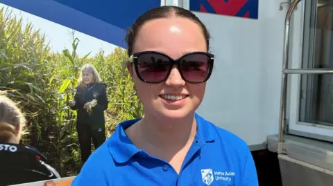 A young woman with brown hair tied back in a ponytail smiles. She wears large dark sunglasses and a blue polo shirt with a white Harper Adams University logo on the shoulder.
