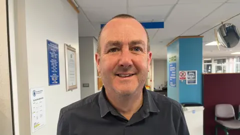 Ian looking into the camera and smiling; he's wearing a black collared shirt. Behind him you can see part of a waiting area at the hospital, it's got blue and white walls with one brown chair just visible to the right of the picture. There's various signs on the walls, but they're out of focus so you can't read what they say. 