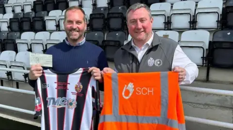 Spennymoor Town FC Gary Finley wearing a navy jumper holds a cheque and a black and white striped football shirt up, as he stands next to a fan, who is wearing a grey gilet with a football emblem on it, over a white shirt. He is holding up an orange hi-vis jacket. Both are standing on a football pitch on a sunny day. Black and white seats are behind them.