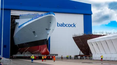 Babcock International The white prow of HMS Venturer emerges on a trolley from a large hangar-style building with the word "babcock" on the side. Workers in yellow and orange overalls and jackets are standing in front of the ship.
