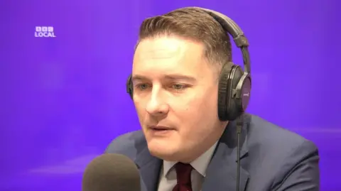 A picture of Wes Streeting wearing headphones and speaking into a microphone. There is a purple background, that says "BBC local" on it.