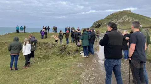 A large mass of people standing on green cliffs looking out towards the sea with the Rumps beyond