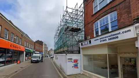 Businesses and a van can be seen on a street in a town. On the right is an area surrounded by boarding and scaffolding.