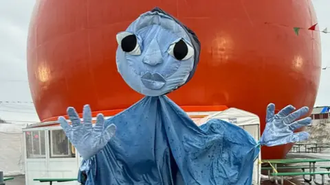 A large blue puppet with a round face draped in a blue robe in front of an orange sphere. The feet of three puppeteers are visible. 