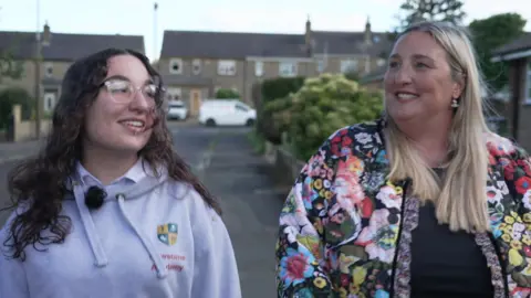 Mary is on the left of the image and has dark brown curly hair and is wearing glasses. Lydia is on the right of the image and has long blonde hair and is wearing a multi-coloured jacket