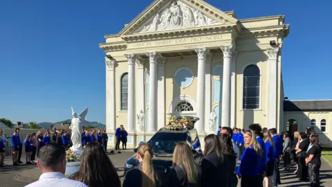 A cream church building with 'Sancta Maria Ora Pro Nobis' on the entrance, held up by four long pillars and in between the pillars a white arch and brown door with two white statues on either side between the pillars. Then in front of the church is another white statue with wings and flowers laid in front. To the right is a black hearse with the words 'EMMANUEL' in white flowers and flowers above on the roof. Students in uniform line up either side of the hearse.