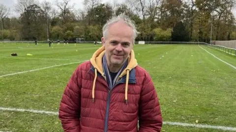 Newport IW FC Paul Rushton poses at the ground of Newport County AFC, leaning against a metal barrier with the pitch in the background. He has grey hair and is wearing a red padded jacket over a yellow hoodie. Trees can be seen in the background beyond the pitch