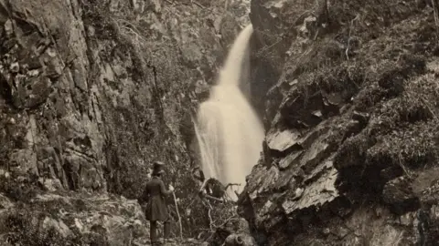 National Trust A black and white image of Aira Force with a man wearing a top hat and long coat standing in front of it, holding a walking stick.