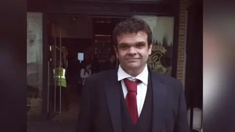 Clare Pounder Jamie Sample, a man with dark hair in a suit and red tie.