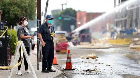 Getty Images Two local residents in masks look on as firefighters continue to work on the fire at the storage facility